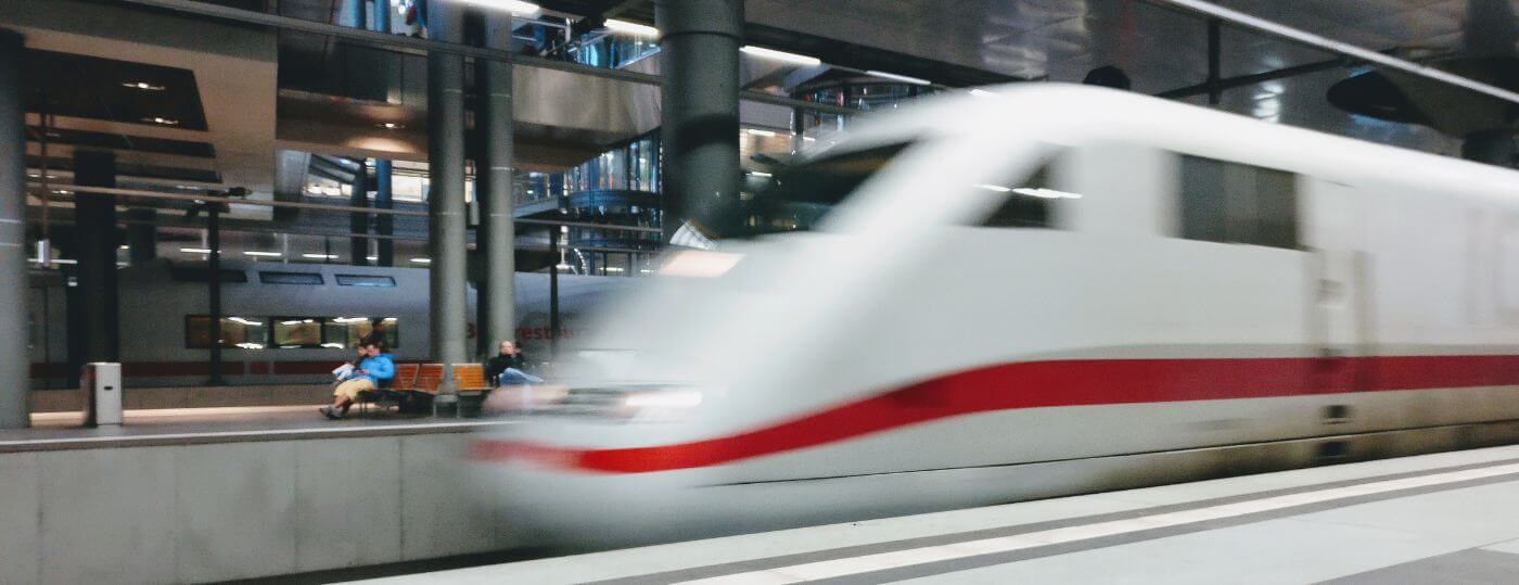 Weißer ICE mit rotem Streifen fährt mit Bewegungsunschärfe durch einen modernen Bahnhof, im Hintergrund sitzen Reisende auf Bänken und Bahnsteig.