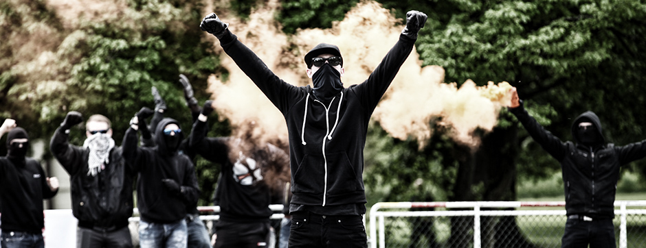 Vermummte Ultras mit einer Rauchbombe im Hintergrund.